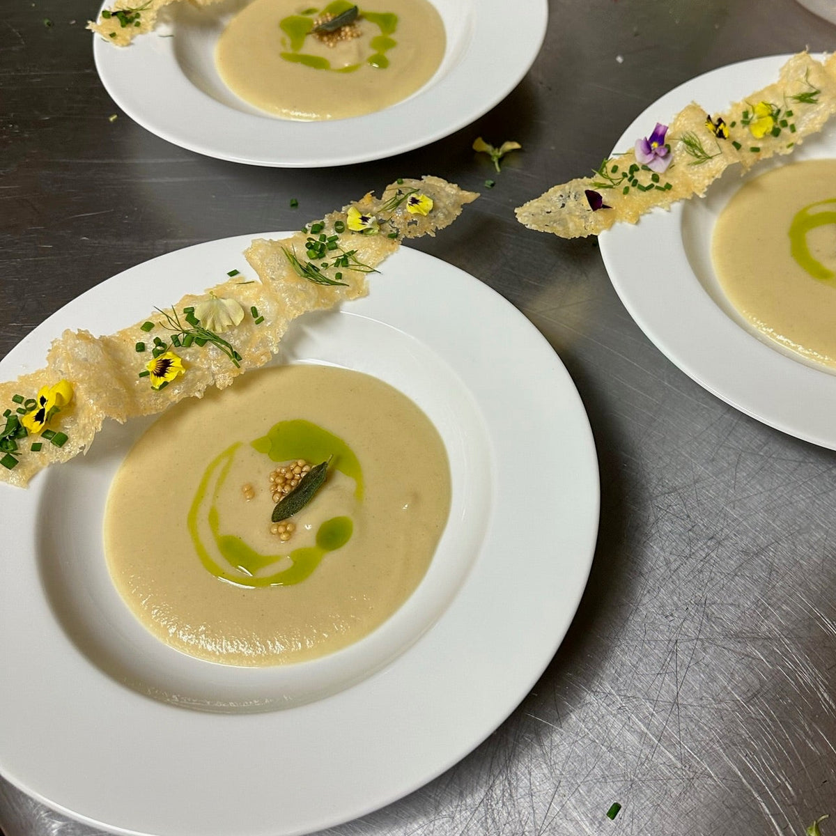 Three white plates of creamy yellow soup garnished with a green oil swirl, herb, and edible flower, each served with a long, crispy cracker strip on a stainless steel kitchen counter.